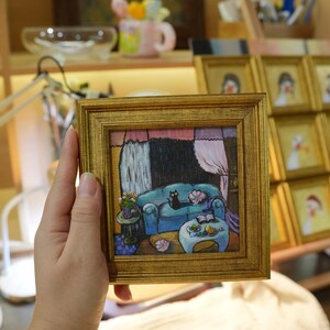 May include: A small, square painting in a gold frame depicts a cozy living room scene. A black cat rests on a blue sofa. The artwork features a table, curtains, and a lamp. Other framed paintings are visible in the background.