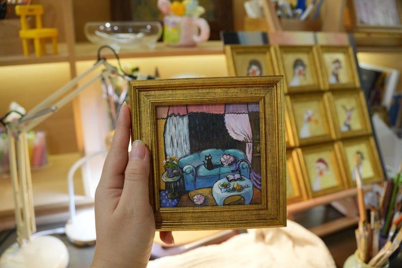 May include: A small, square painting in a gold frame depicts a cozy living room scene. A black cat rests on a blue sofa. The artwork features a table, curtains, and a lamp. Other framed paintings are visible in the background.