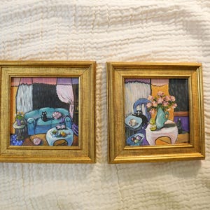 May include: Two framed paintings with gold frames. Each painting depicts a room interior with a cat. One shows a blue sofa, the other a table with flowers. The backgrounds have pink, purple, and black elements.