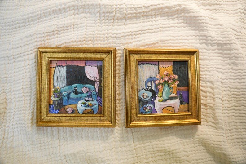May include: Two framed paintings with gold frames. Each painting depicts a room interior with a cat. One shows a blue sofa, the other a table with flowers. The backgrounds have pink, purple, and black elements.