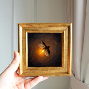 Puede incluir: Una pintura al óleo cuadrada en un marco dorado que representa una luciérnaga con el abdomen brillante sobre un fondo oscuro y arremolinado. El insecto está centrado, con las alas extendidas y una luz cálida y circular emanando de su cuerpo.