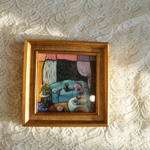 May include: A framed artwork depicts a cozy interior scene. A teal sofa holds a black cat and a sleeping figure. A small table with flowers sits nearby. The artwork is in a gold frame, set against a white lace background.