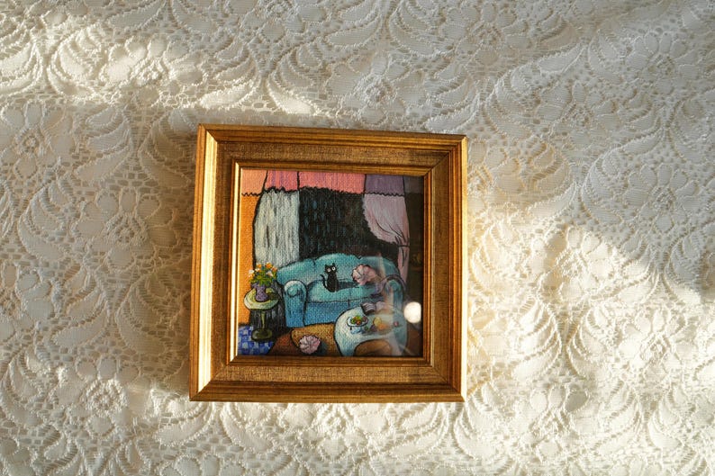 May include: A framed artwork depicts a cozy interior scene. A teal sofa holds a black cat and a sleeping figure. A small table with flowers sits nearby. The artwork is in a gold frame, set against a white lace background.