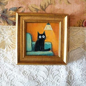 May include: Square framed painting of a black cat with large white eyes, sitting on a teal armchair under a lit lamp. The background is orange, and the frame is gold. The painting is on a floral background.