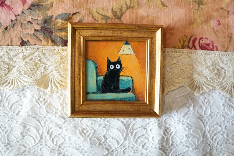 May include: Square framed painting of a black cat with large white eyes, sitting on a teal armchair under a lit lamp. The background is orange, and the frame is gold. The painting is on a floral background.