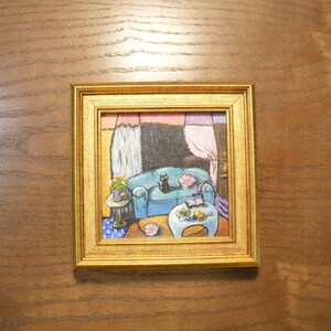 May include: A framed painting depicts a cozy living room scene. A blue sofa with a black cat is the focal point. A small table with flowers and a coffee table with fruit are also visible. The frame is gold.