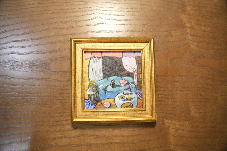 May include: A framed painting depicts a cozy living room scene. A blue sofa with a black cat is the focal point. A small table with flowers and a coffee table with fruit are also visible. The frame is gold.
