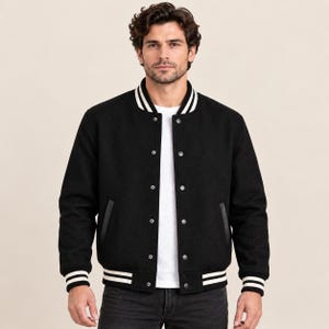 Könnte beinhalten: Eine schwarze Collegejacke mit weiß gestreiftem Rippstrickkragen, Bündchen und Saum. Die Jacke verfügt über eine Druckknopfleiste, zwei Seitentaschen mit schwarzer Einfassung und lange Ärmel. Sie wird über einem weißen T-Shirt getragen.