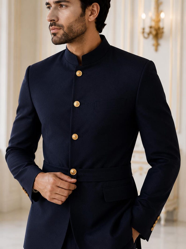 May include: A dark navy blue blazer featuring a high mandarin collar, long sleeves, and a front button closure with four polished gold-tone buttons. The jacket includes a matching integrated waist belt and two flap pockets on the lower front.