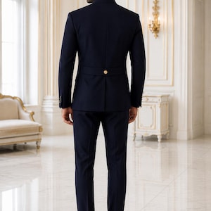 May include: A dark navy blue suit featuring a jacket with a mandarin collar, long sleeves, and a horizontal back strap detail secured with a single gold-toned button. The matching trousers are slim-fit, paired with black leather dress shoes.