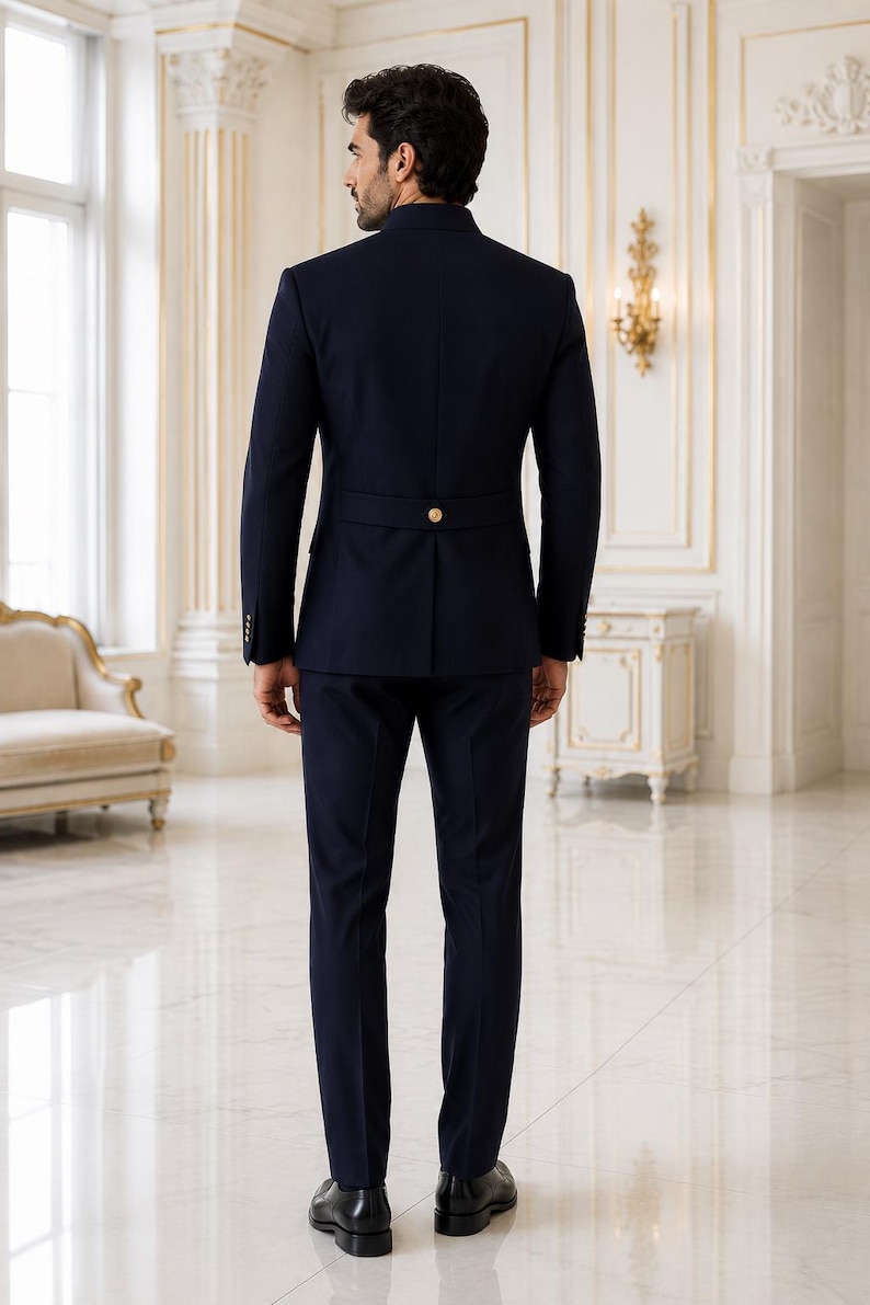 May include: A dark navy blue suit featuring a jacket with a mandarin collar, long sleeves, and a horizontal back strap detail secured with a single gold-toned button. The matching trousers are slim-fit, paired with black leather dress shoes.