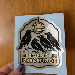 May include: A sticker with a light blue background featuring four black crows and a disco ball. The text "MURDER ON THE DANCEFLOOR" is displayed in a gold font. The sticker has a silver border.
