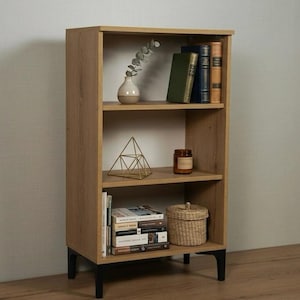 May include: A light brown wooden bookcase with three shelves. The shelves hold books, a small vase with greenery, a geometric gold decoration, a candle, and a woven basket. The bookcase has black legs.