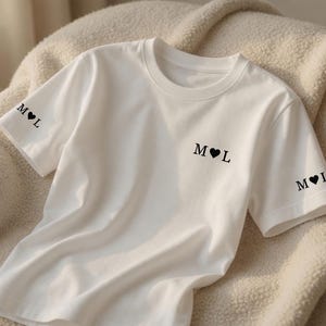 Könnte beinhalten: Weißes T-Shirt mit Rundhalsausschnitt. Auf dem Shirt sind die Initialen "M" und "L" mit einem Herzsymbol in Schwarz auf der Brust und an den Ärmeln aufgedruckt. Das Shirt besteht aus einem weichen, glatten Stoff.