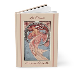 May include: A hardcover book with a light beige cover featuring the text La Danse and Élégance Éternelle Art Nouveau Paris. The central illustration depicts a figure with long flowing red hair draped in pink fabric against a circular frame.