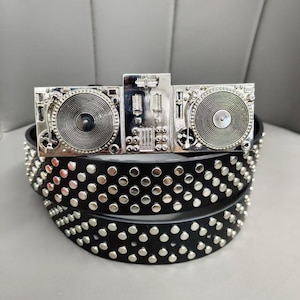 Könnte beinhalten: Ein schwarzer Gürtel mit einer silberfarbenen Schnalle in Form eines DJ-Mixers mit zwei Plattenspielern. Der Gürtelriemen ist mit drei horizontalen Reihen von gleichmäßig verteilten, runden, silberfarbenen Metallnieten verziert.