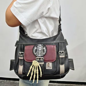 Peut inclure: Un sac hobo en similicuir noir avec une poche avant bordeaux, des surpiqûres blanches et des boucles argentées. Un écusson circulaire représente un squelette et une breloque en forme de main de squelette crème pend du rabat avant.