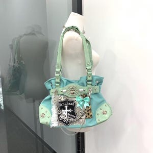Borsa tote in tela verde menta stile Y2K, toppa a croce con teschio punk, inserti in pizzo leopardato e scozzese, cinturino in pelle di coccodrillo e decorazione a cuore.