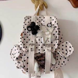 May include: A cream-coloured backpack with black polka dots, featuring two front pockets, white canvas straps with metal buckles, a black fabric flower, a glittery star-shaped charm, a silver chain, and a small circular pin with a skull graphic.