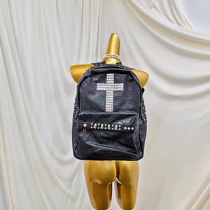 Peut inclure: Un sac à dos noir avec un motif de crânes en relief sur toute la surface. L'avant présente une grande croix cloutée argentée et une sangle horizontale ornée de petits crânes et de clous ronds argentés au-dessus de la poche avant.
