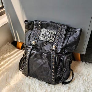 May include: A black textured leather backpack featuring a large rectangular silver-toned metal buckle with intricate embossed detailing on the front flap. The bag includes silver studs, two spider-shaped ornaments with pearl accents, and side pockets.