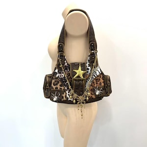 Peut inclure: Un sac à épaule marron avec un motif léopard, un emblème central en forme d'étoile dorée et une breloque en chaîne dorée avec des clochettes. Le sac comprend deux poches latérales, des araignées argentées avec des perles blanches et une boucle.