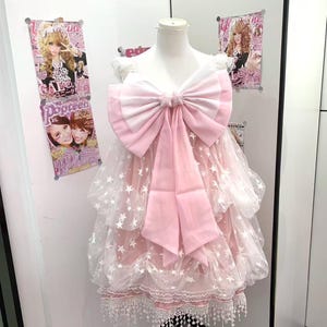 May include: A sleeveless dress featuring a large pink and white bow at the chest, layered tulle fabric with a white star pattern, and a white lace trim along the hem. The garment is displayed on a mannequin, showcasing a tiered, voluminous silhouette.