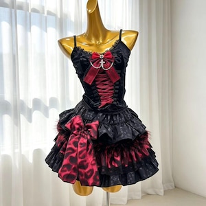 May include: A black sleeveless dress featuring a red lace-up corset bodice with a central red bow and silver cross charm. The skirt is layered with ruffled fabric, a black star pattern, and a large red and black leopard print bow at the side.