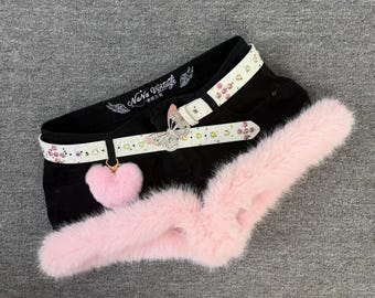 May include: Black denim shorts featuring a white belt with rhinestones and a silver butterfly buckle. A pink faux fur trim lines the bottom edge, and a pink heart-shaped pom-pom charm hangs from the belt. The inner waistband displays the text NaNa Vintage.