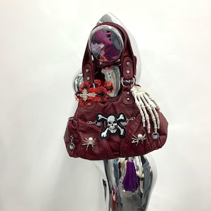 Op de afbeelding: Een donkerrode handtas met een centrale schedel- en gekruiste beenderen-patch, een witte skeletthand, twee zilveren spinnenbedels en een rood kanten kruis. De tas heeft twee handvatten met zilverkleurige metalen hardware en klinknagels.