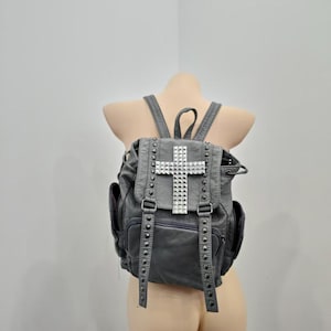 May include: A charcoal grey backpack featuring a prominent silver-toned studded cross on the front flap. The bag includes two vertical studded straps, a drawstring closure, side pockets, and adjustable shoulder straps with a textured finish.