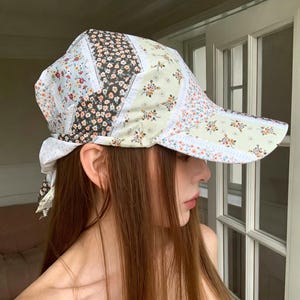May include: A baseball cap featuring a patchwork design of various floral patterns in shades of light green, white, orange, and black. The cap has a structured brim and a fabric tie closure at the back, constructed from multiple panels of printed cotton fabric.