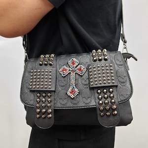 May include: A black shoulder bag featuring an embossed skull pattern, adorned with metallic studs on two front flaps and a central cross emblem encrusted with red and blue rhinestones. The bag includes a black shoulder strap and metal hardware.
