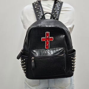 Könnte beinhalten: Ein schwarzer Rucksack aus Kunstleder mit Krokodilleder-Textur und einem auffälligen roten Kristallkreuz auf der Vordertasche. Der Rucksack ist mit silberfarbenen Metallnieten an den Seitentaschen und Schultergurten sowie einem Tragegriff versehen.