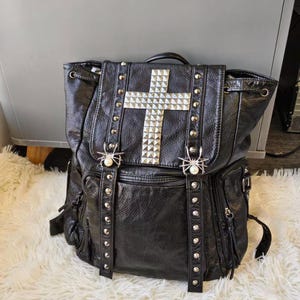 May include: A black textured backpack featuring a large silver pyramid-studded cross on the front flap. The bag includes two vertical studded straps, each adorned with a silver spider ornament featuring a white pearl-like bead at the centre.