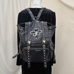 May include: A black leather backpack featuring a silver-toned cobra emblem on the flap, two spider-shaped ornaments with white pearls, and vertical rows of metal studs. The bag includes a drawstring closure, adjustable straps, and a front pocket.