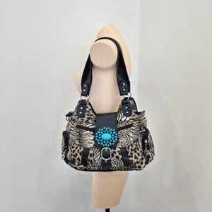 Peut inclure: Un sac à bandoulière avec un motif léopard noir et beige et des garnitures en similicuir noir. Le rabat avant est fermé par une boucle argentée et orné d'un grand médaillon ovale en perles turquoise. Il possède deux anses noires.
