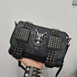 May include: A black shoulder bag featuring an embossed skull pattern across the surface. The front flap is decorated with a central silver-toned dragon head ornament with red eyes, flanked by two rectangular panels covered in silver metal studs.