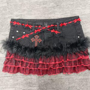 Puede incluir: Falda vaquera negra con volantes de encaje rojo y ribete de plumas negras. La falda presenta lazos rojos y un diseño de cruz. Un artículo de moda de inspiración gótica.