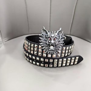 Peut inclure: Une ceinture en cuir noir ornée de deux rangées de clous métalliques carrés argentés. La boucle de ceinture est une grande tête de dragon argentée détaillée avec des cornes, une gueule ouverte et de petits yeux rouges brillants incrustés.