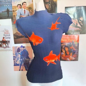 May include: A navy blue, short-sleeved top with a high neckline. The top has a textured, pleated design and is decorated with three bright orange goldfish. The garment is displayed on a mannequin.