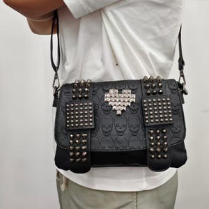 May include: A black crossbody bag featuring an embossed skull pattern across the surface. The bag is decorated with silver-toned metal pyramid studs arranged in rectangular patches and a central heart-shaped design made of metallic studs.