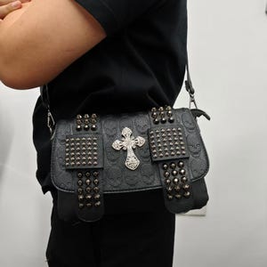 May include: A black crossbody bag featuring an embossed skull pattern on the leather-like material. The bag is decorated with silver-toned metal studs on the front flap and a prominent, ornate silver-toned cross emblem centered on the front.