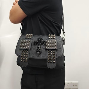 May include: A black crossbody bag featuring an embossed skull pattern on the leather-like material. The flap is decorated with two vertical straps covered in dark metallic studs and a central black beaded cross embellishment with a shoulder strap.