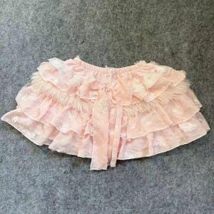 Peut inclure: Une jupe rose clair composée de plusieurs étages de tissu à volants avec un motif floral brodé ton sur ton. Le vêtement présente une taille élastique et un panneau central vertical de tissu qui tombe en cascade sur les couches avant.