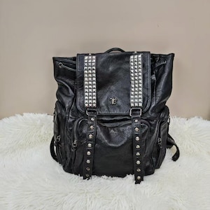 May include: A black backpack featuring a textured finish, two vertical straps adorned with silver-toned pyramid studs, and a central metal emblem. The bag includes a flap closure, side pockets with zippers, and a drawstring top for secure storage.