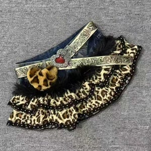 May include: A pair of dark blue denim shorts featuring a tiered leopard print ruffle hem with black lace trim. The shorts include a belt with an engraved floral pattern, a central heart-shaped buckle, and a fuzzy leopard print heart charm.