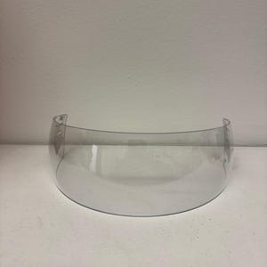 以下が含まれることがあります： 透明で湾曲したバイザー。ヘルメット用と思われる。透明なプラスチック製。滑らかで丸みを帯びた形状で、顔を保護するように設計されている。素材は軽量で耐久性があるように見える。