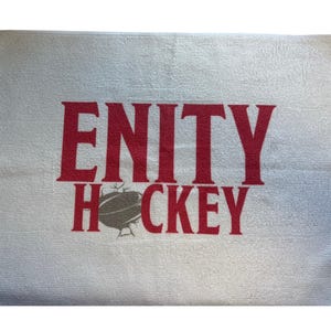 以下が含まれることがあります： 白いマイクロファイバータオル。赤いブロック体で「ENITY HOCKEY」の文字がプリントされています。「ENITY」の下には灰色のホッケーパックのグラフィックがあります。小さなタグが右上隅に付いています。