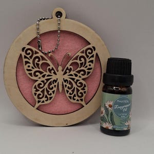 May include: A wooden butterfly ornament with a pink backing, alongside a small bottle of fragrance oil. The ornament has a chain. The fragrance oil bottle features a green label with white daisies and the text "PHATOIL Fragrance Oil La Vie Est Belle".